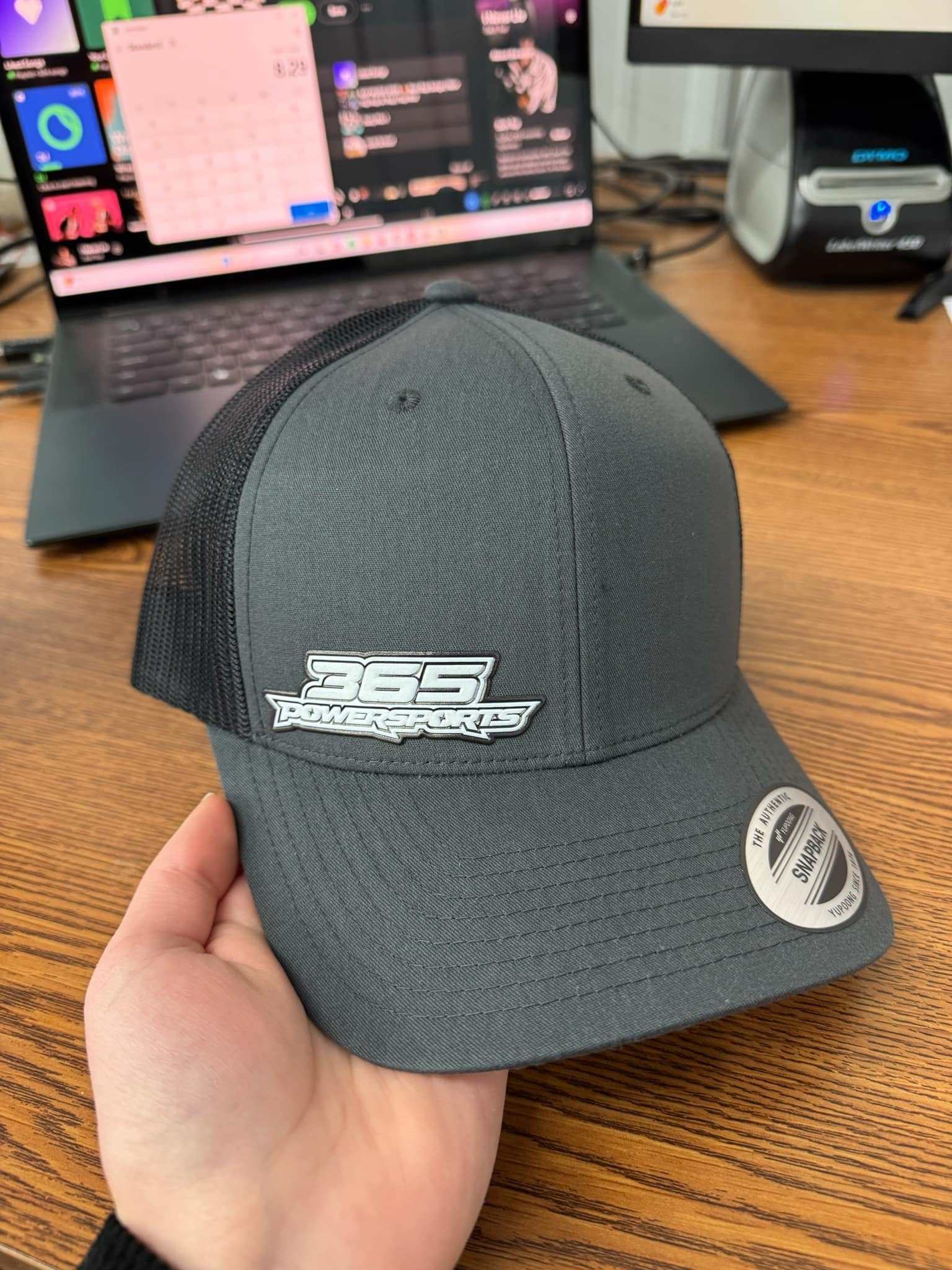 365 Retro Trucker flex patch hat in black with mesh back held over wooden desk with laptop in background